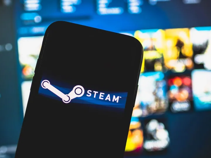 A smartphone displaying the Steam logo in focus, with a blurred background featuring what appears to be a monitor with various game graphics.
